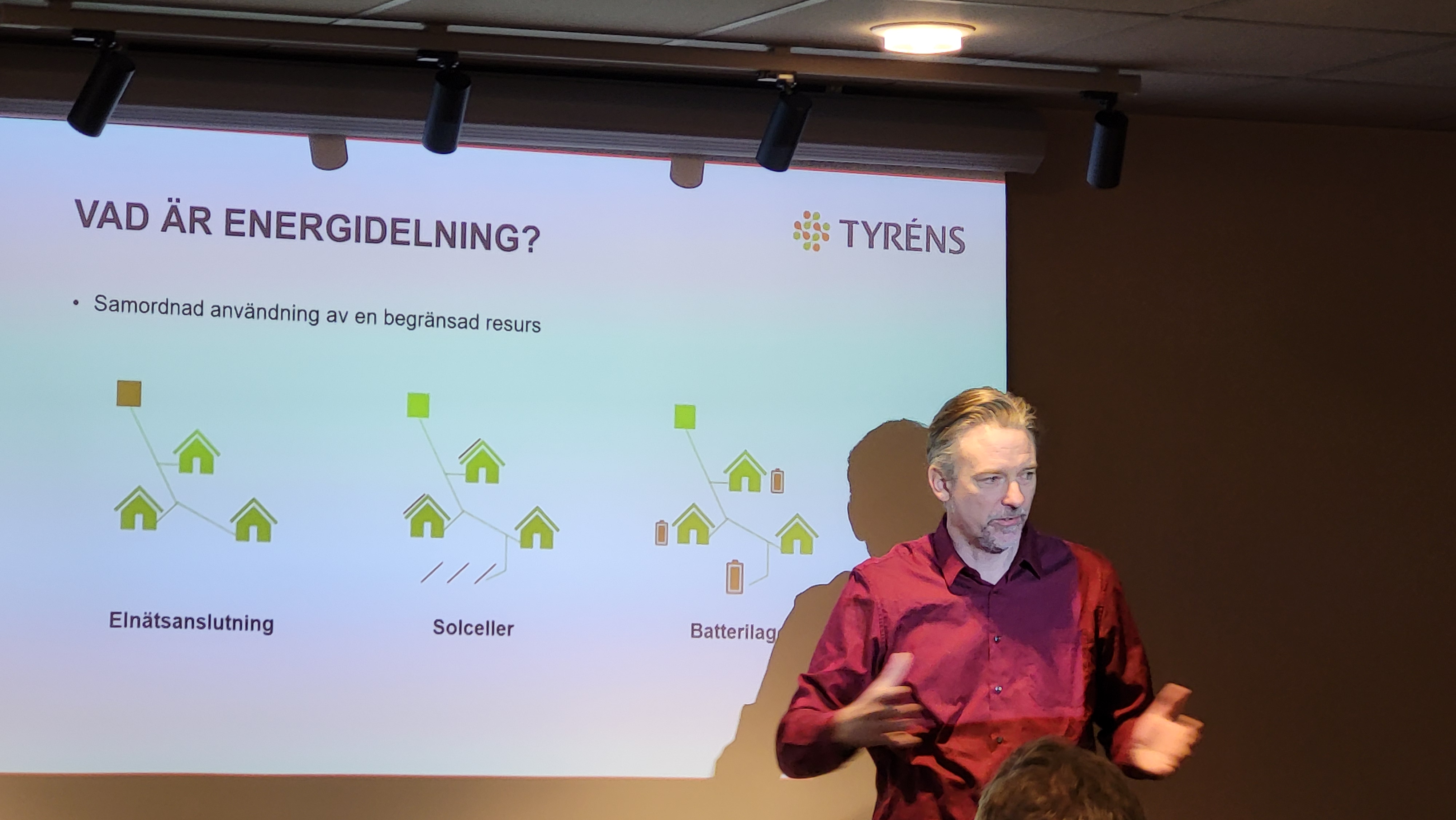 En gråhårig man med skägg, i vinröd skjorta, står framför en skärm med en bild som förklarar Vad är energidelning. Illustrationen visar grupper med hus som delar energi mellan varandra.