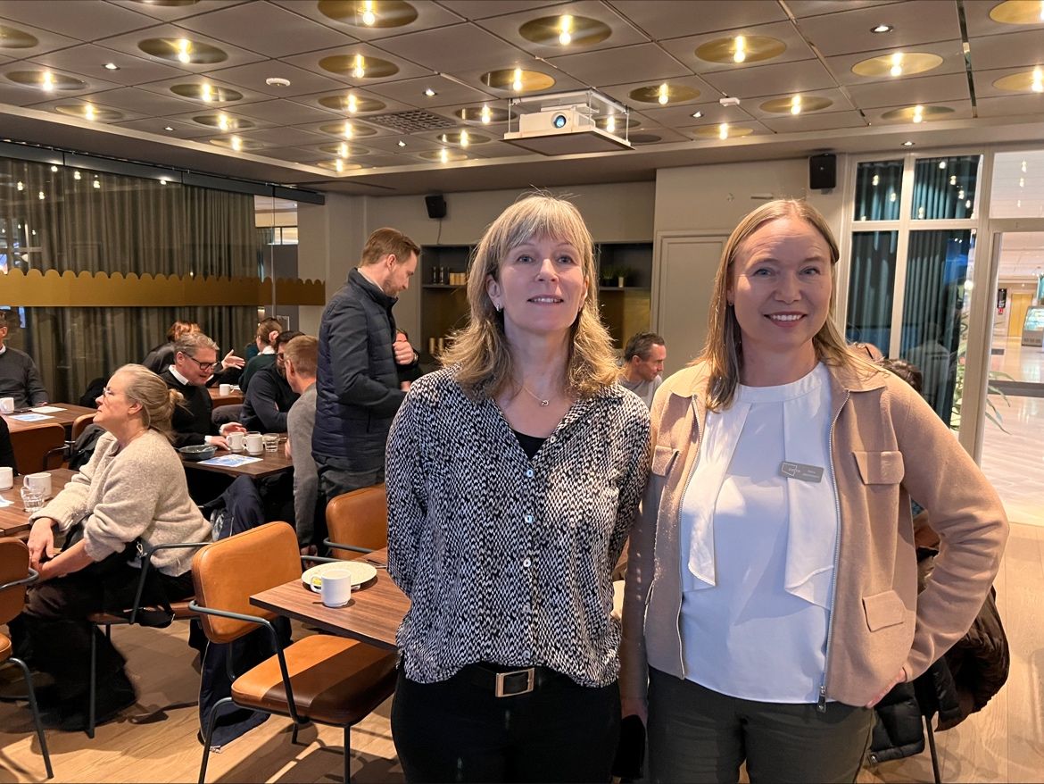 Två kvinnor tittar in i kameran, de står bredvid varandra och ler. I bakgrunden syns människor som minglar och äter frukost tillsammans