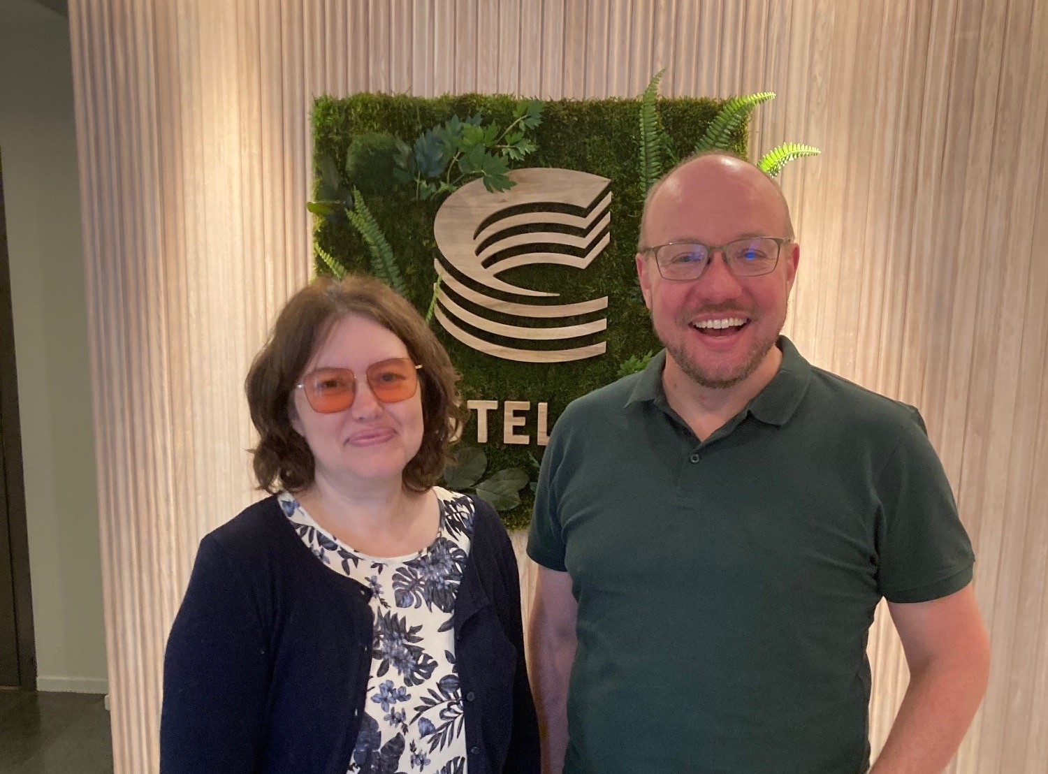 Bild på en kvinna i blå och vit klänning och en man i grön tröja och svarta byxor. De står i ett rum med castellums logga i guld på grön bakgrund.  