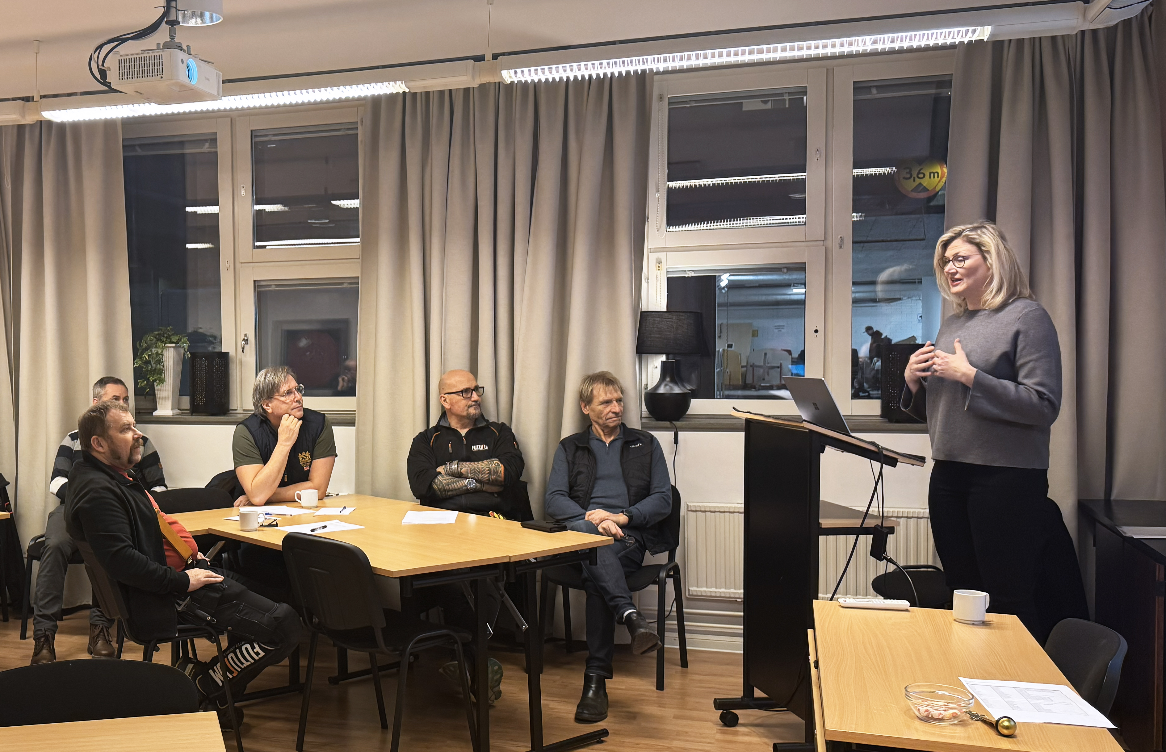 ett rum med människor som sitter  och tittar på en stor skärm med en föreläsare som pratar