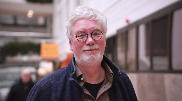 En vithårig man med skägg och glasögon står framför några hus.