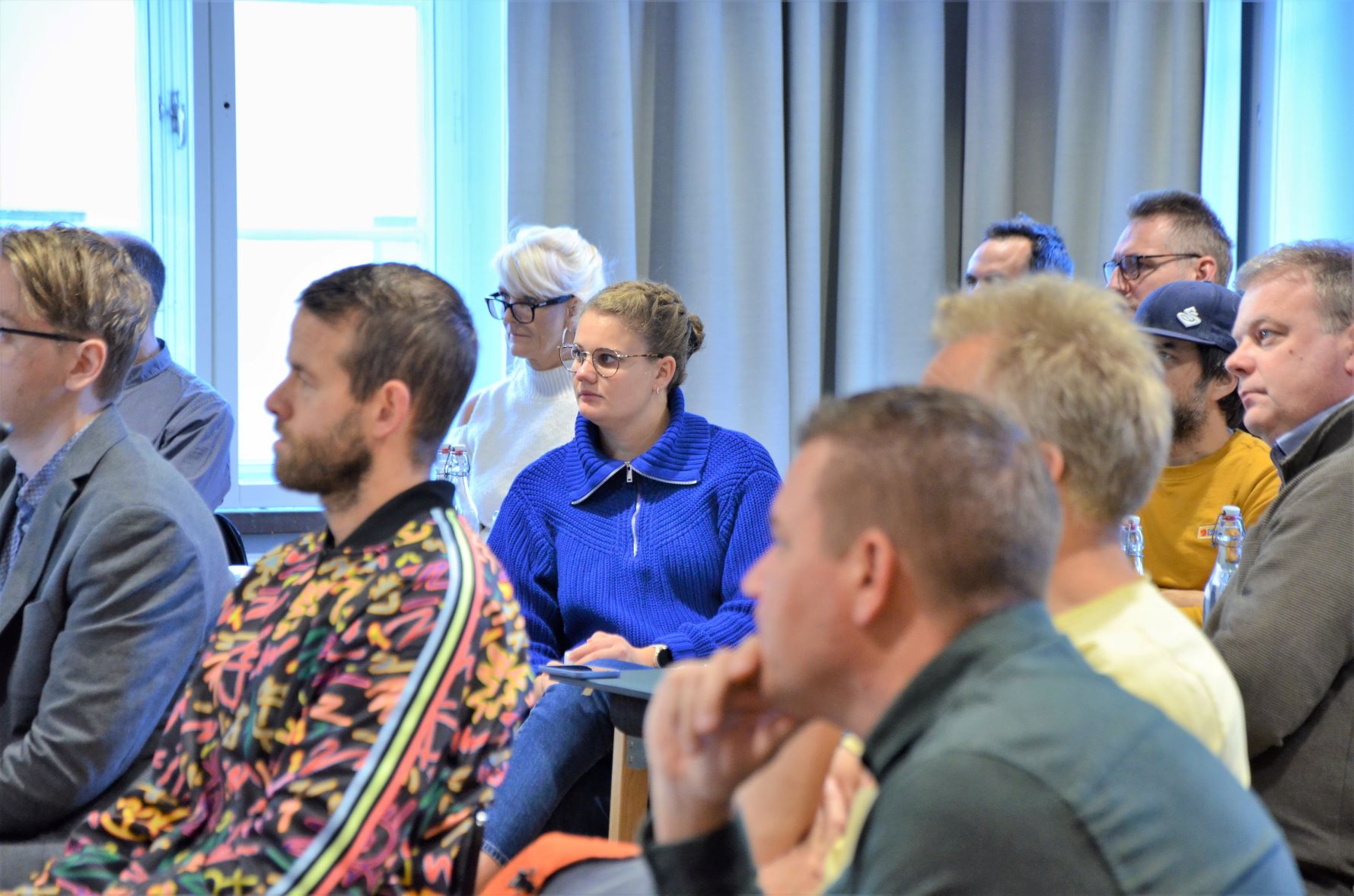 Bild på åhörare, en kvinna med brunt hår, glasögon och blå tröja lyssnar på föreläsaren, runt henne sitter flera andra som lyssnar