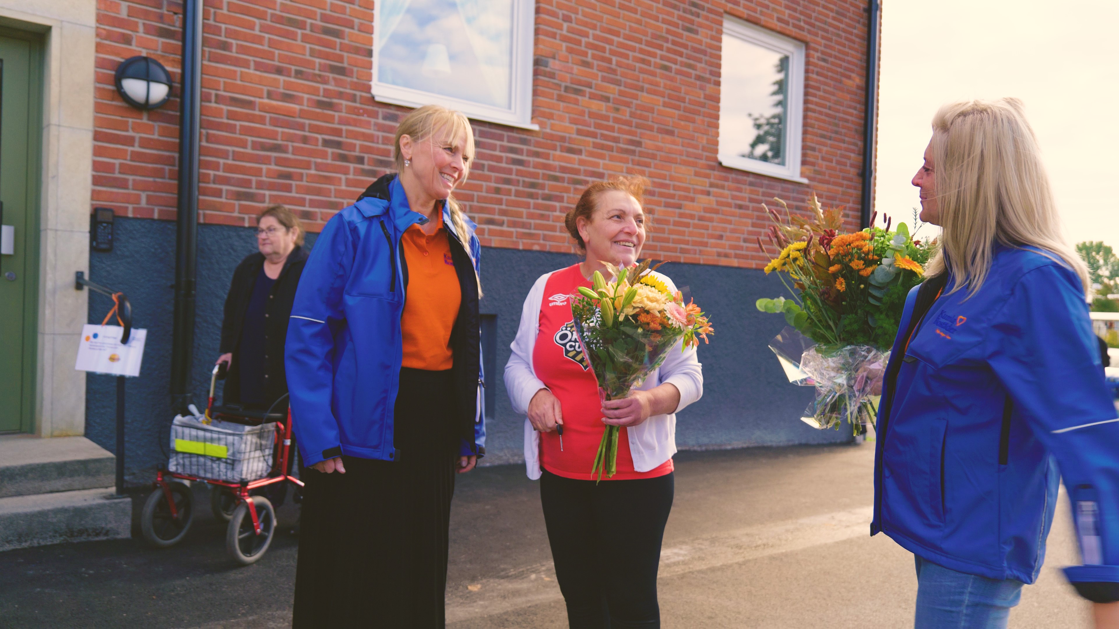 Två pensionärer, en man och en kvinna, står och ler med en blombukett i handen. Mitt emot står Jaana Ekeblom och ler, också med en blombukett i handen. 