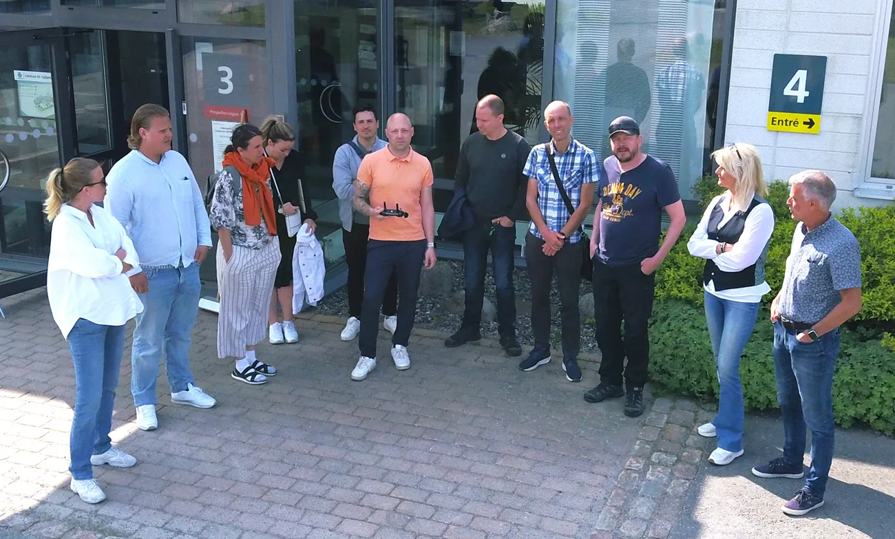 Bilden är tagen ifrån en drönare. Ett gäng personer står framför en byggnad och tittar upp mot luften, det är sommar och många ler.