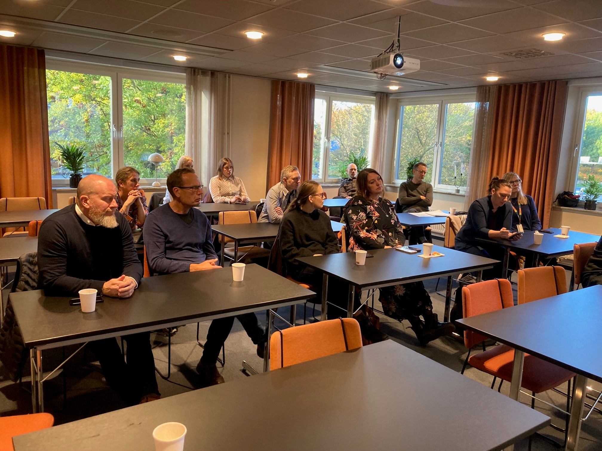Bild på en liten föreläsningssal med orangea gardinger och svarta bord. De står i klassuppställning och det är blandat med män och kvinnor som sitter och lyssnar. ca 12 personer på bilden