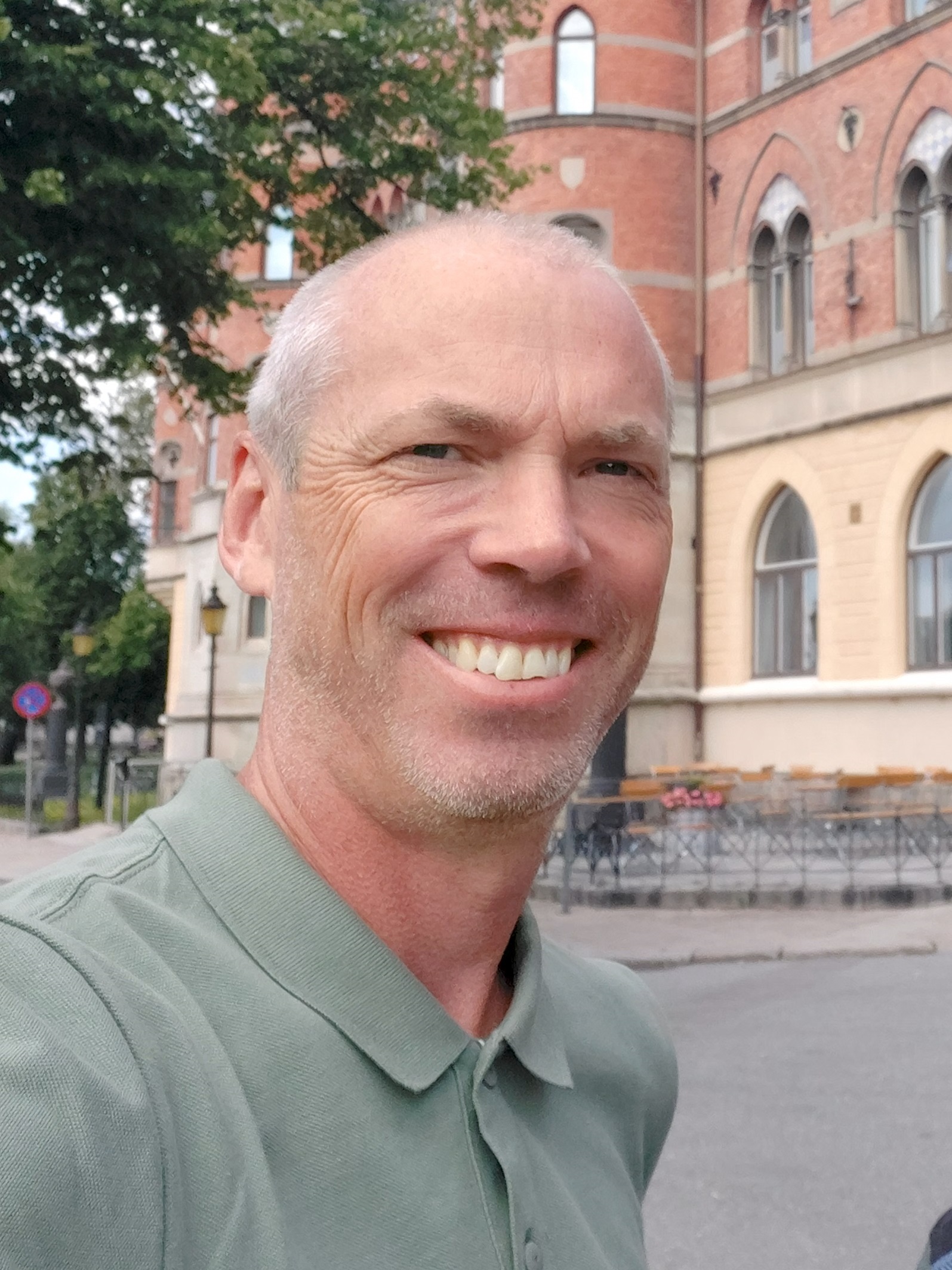 Bild på en man i närbild med en borgliknade hus i bakgrunden. Han har en grå tröja på sig. 