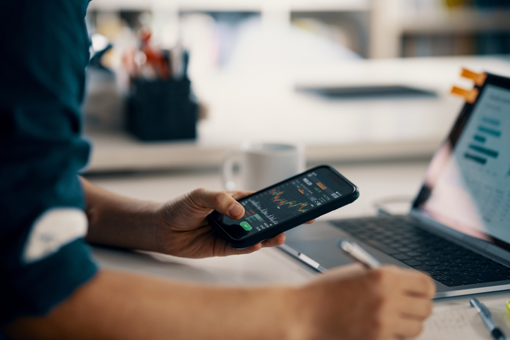Person håller i en smartphone med grafer på, framför sig har han en dator
