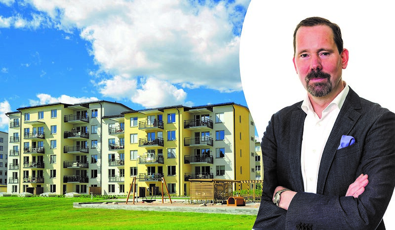 En mörkårig man med kort skägg i grå kavaj på högersida och vänstra sidan är det lägenhetshus i gul och vitt med en lekplasts i förgrunden och det är en fin dag med blå himmel och några moln