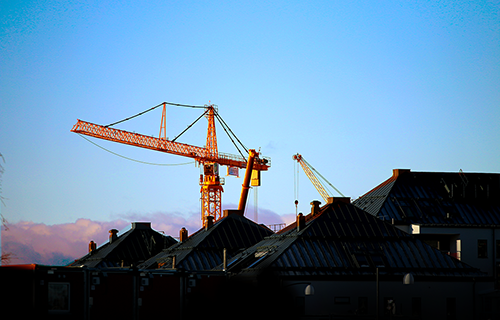 hus i förgrunden som är skuggade med en lyftkran i gul som solen lyster på med en blå himmel bakom