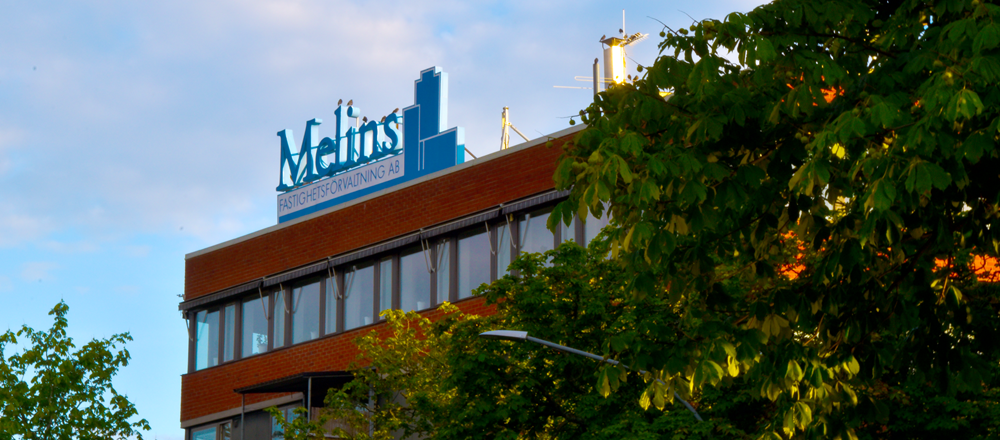 Melins blå logotyp på taket till ett hus i rött med många fönster. 50-60tals stil med ett träd i förgrunden på högersida