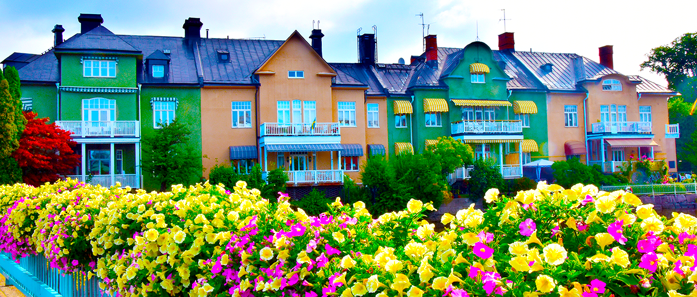 Fyra hus, vartannat grönt och vartannat beige, i olika utföranden och detaljer. Framför husen finns en mängd gula och lila blommor i blomlådor som satts fast i ett blått staket.
